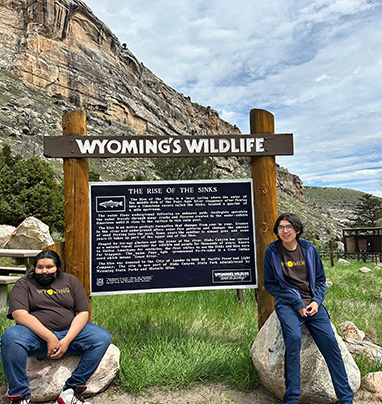 two boys sitting on rocks under a sign that reads Wyoming's Wildlife with information too small to read about the Rise of the Sinks next to a picture of a fish