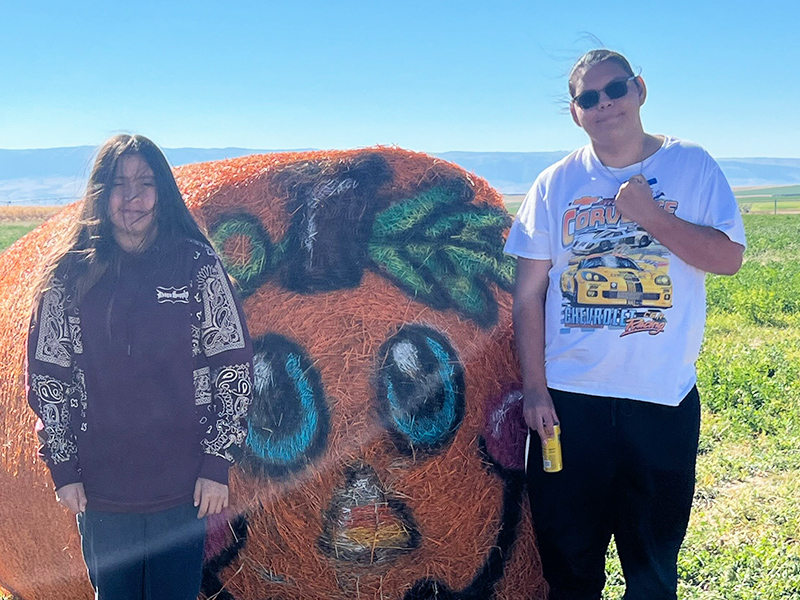 Two students outside next to a huge pumpkin made of straw
